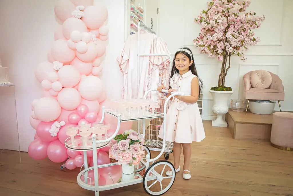 A young girl in a pale pink dress stands smiling beside a white cart with drinks, surrounded by pink balloons, flowers, and a flowering tree—perfect for capturing memories with a kids birthday party photographer in this stylish, pastel-decorated room.