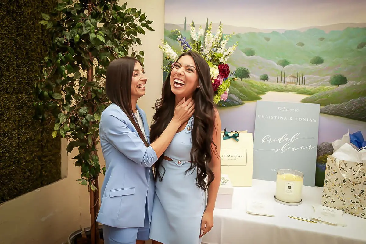 Two women in light blue outfits laugh together at a bridal shower, standing beside gifts, a candle, and flowers—a joyful moment captured by a queer bridal shower photographer celebrating that love is love. A scenic landscape painting sits in the background.