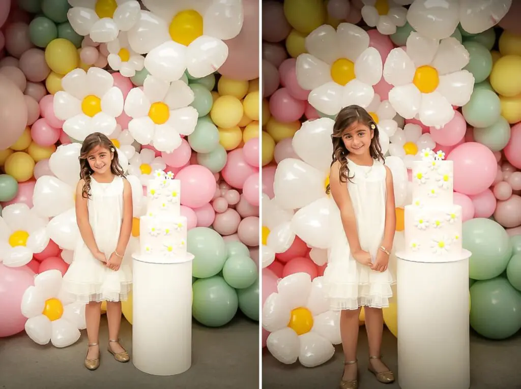 A young girl in a white dress smiles beside a white cake decorated with yellow and white flowers, standing before a colorful balloon wall. Perfect moment captured by a kids birthday party photographer.