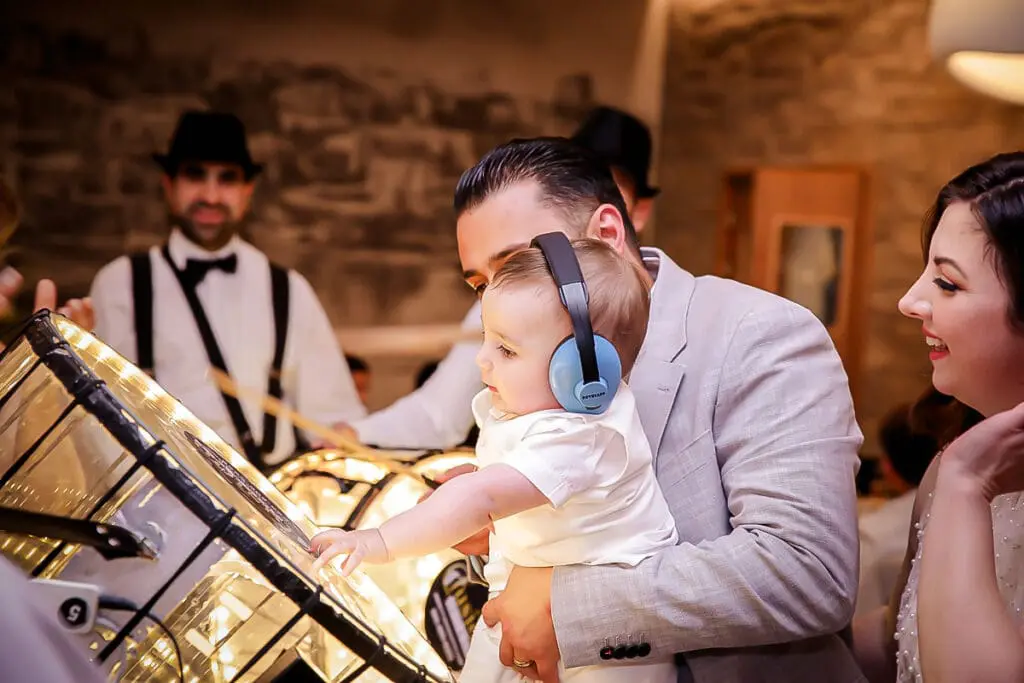 Baby in earmuffs taps a glowing drum while a parent holds them, smiles all around