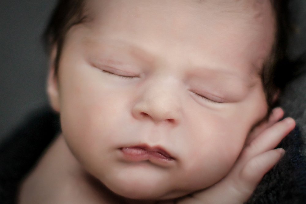 Close up of face and features. Newborn Baby William laying against a gray backdrop, wrapped in a blue newborn stretch.  