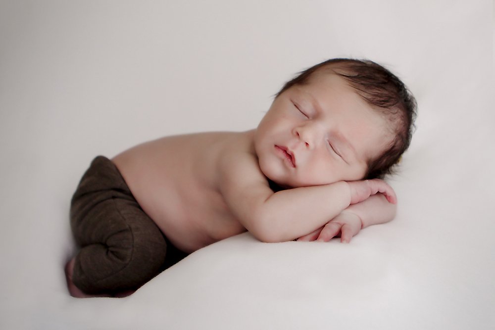 Newborn Baby William laying against a white backdrop,  wearing brown newborn pants sleeping. 
