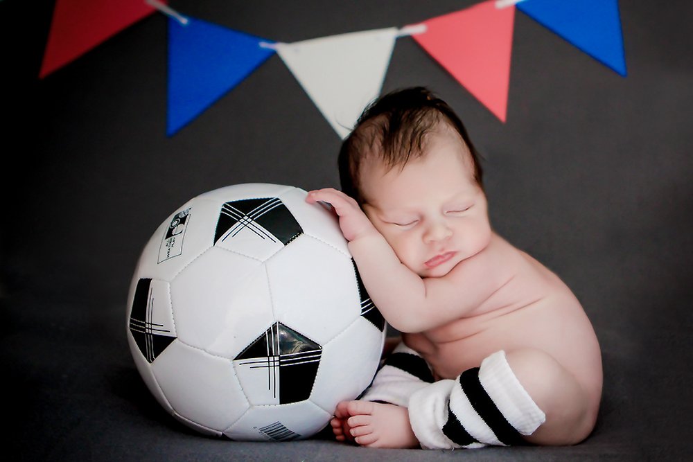 Newborn Baby William laying against a gray backdrop, holding and laying fast asleep on a soccer ball. French Soccer flags are on the background. 