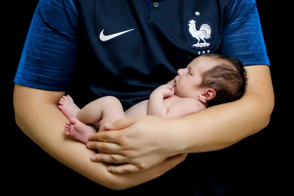 Newborn Baby William in his father's arms, against a black backdrop, fast asleep. His dad is wearing his French Nike Soccer shirt.