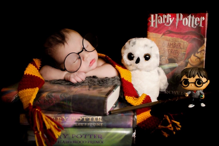 Harry Potter themed newborn photosession. Baby William is laying on a stack of Harry Potter books, holding a wand, wearing a Gryffindor scarf, Harry Potter round glasses, stuffed owl, and a Harry Potter Funko Pop doll used as props.