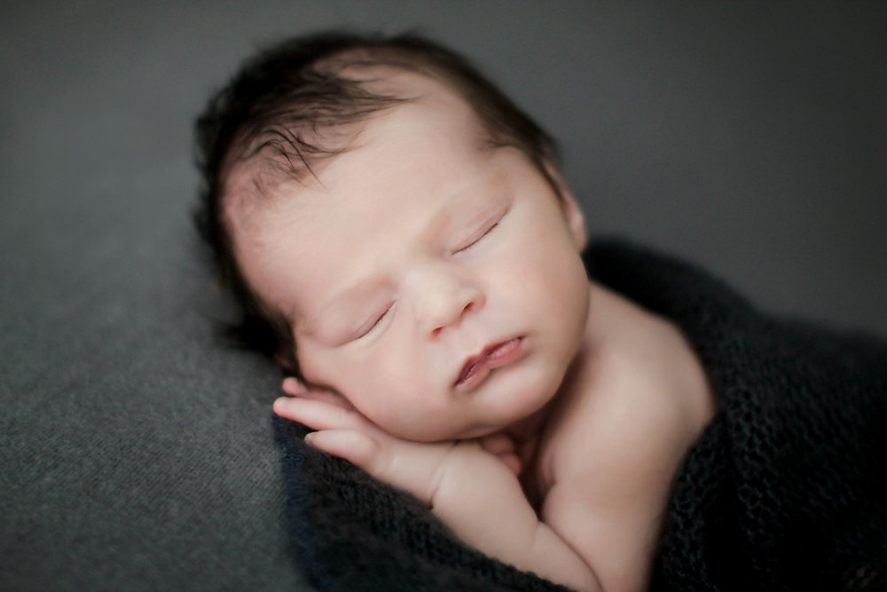 Newborn Baby William laying against a gray backdrop, holding his hands, wrapped in a blue newborn stretch wrap sleeping. 