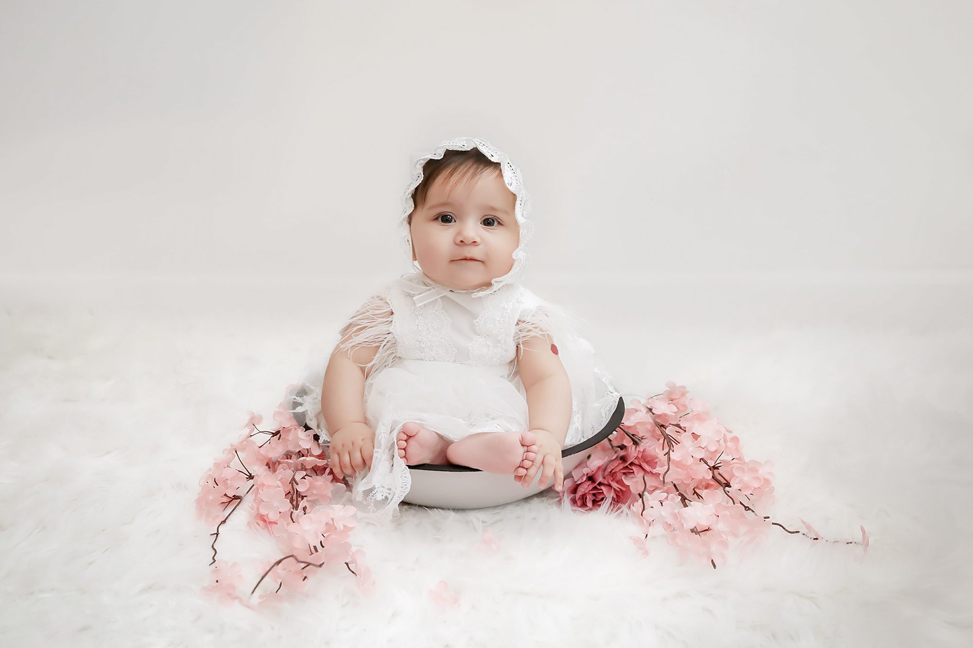 A baby wearing a white bonnet and dress sits in a round bowl on a soft white surface, surrounded by pink artificial flowers, looking at the camera with a calm expression.