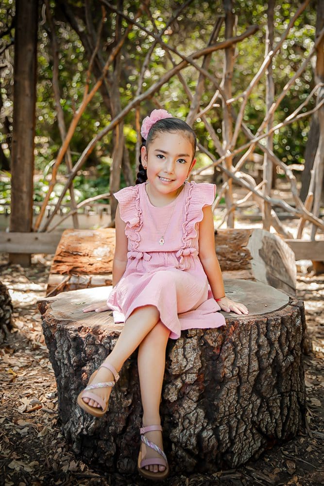 Family-Holiday-Portrait-Session-Descanso-Gardens Smiling girl in a pink dress on a tree stump in a leafy garden