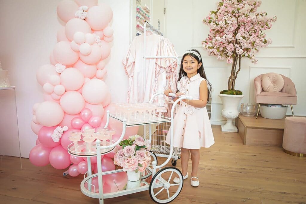 A young girl in a pale pink dress stands smiling beside a white cart with drinks, surrounded by pink balloons, flowers, and a flowering tree—perfect for capturing memories with a kids birthday party photographer in this stylish, pastel-decorated room.