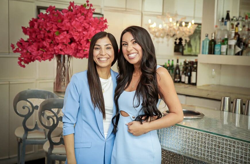Two women in light blue outfits—possibly lesbian brides—smile and pose together in a modern bar or restaurant. Behind them is a large bouquet of bright pink flowers and stylish shelves with bottles and glasses, radiating the spirit of love is love.