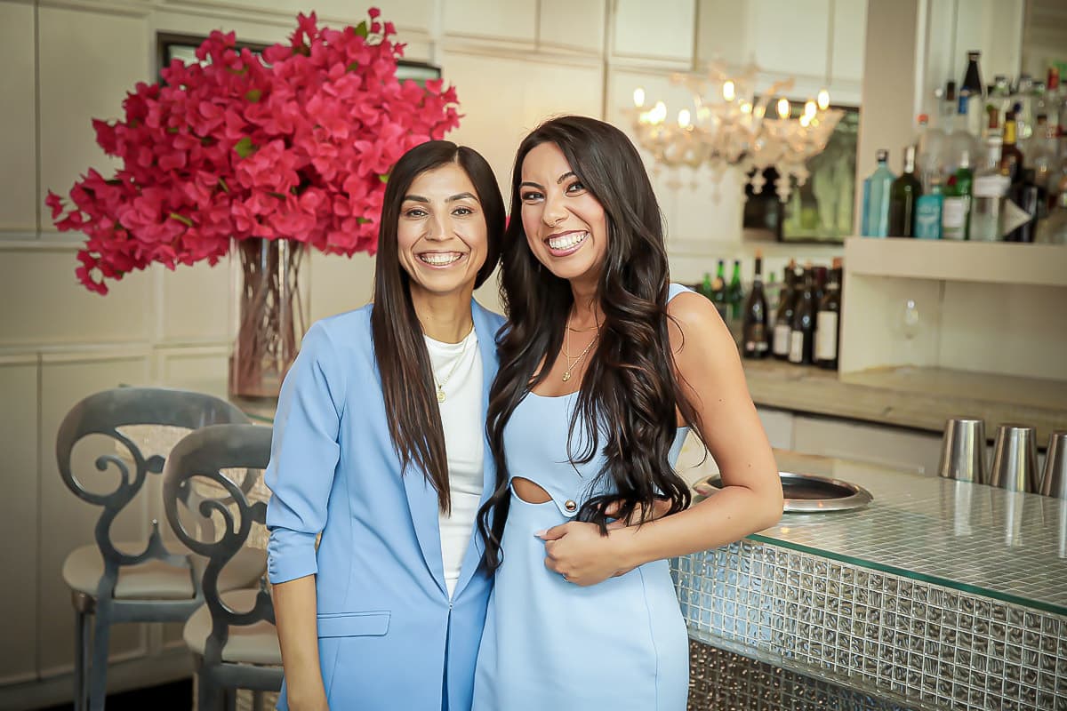 Two women in light blue outfits—possibly lesbian brides—smile and pose together in a modern bar or restaurant. Behind them is a large bouquet of bright pink flowers and stylish shelves with bottles and glasses, radiating the spirit of love is love.