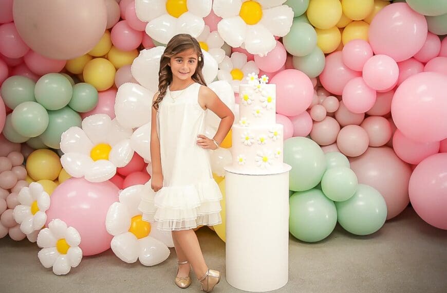 A young girl in a white dress poses beside a white cake decorated with small yellow and white flowers. The background is filled with pastel-colored balloons and large artificial flowers.