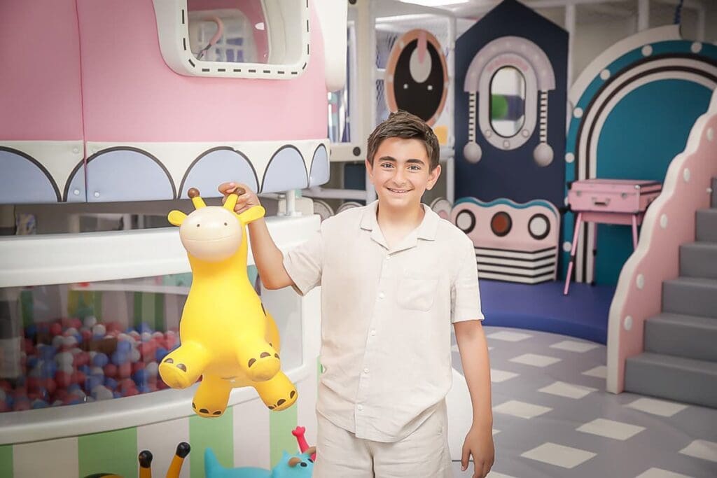 A smiling boy in a white outfit holds a large yellow giraffe toy in a colorful indoor play area, captured perfectly by a kids birthday party photographer, with whimsical decorations and a ball pit in the background.