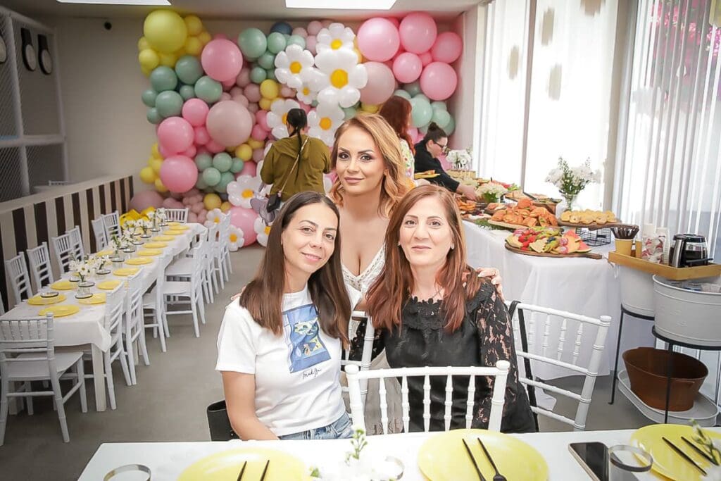 Three women pose and smile in a bright room decorated with pastel balloons and large flower shapes, ready for a kids birthday party. A kids birthday party photographer could capture the cheerful atmosphere, with sunlight streaming through large windows.