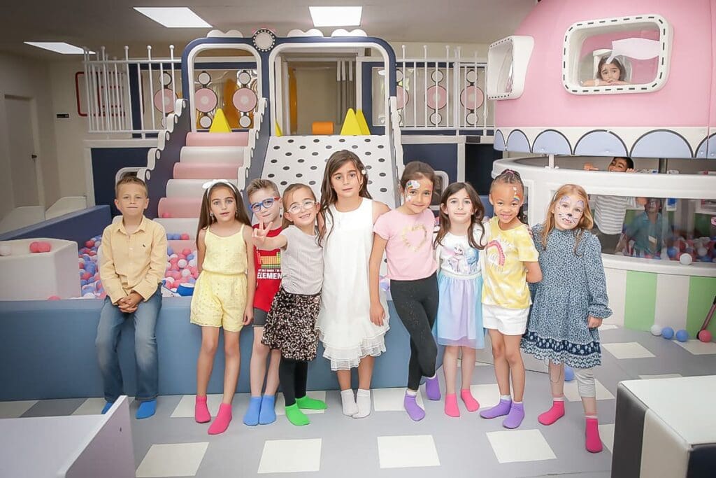 A group of nine children wearing colorful socks stand in a line, smiling at a play area with pastel decor, as a kids birthday party photographer captures the fun by the ball pit and slide, with another child visible through the window.