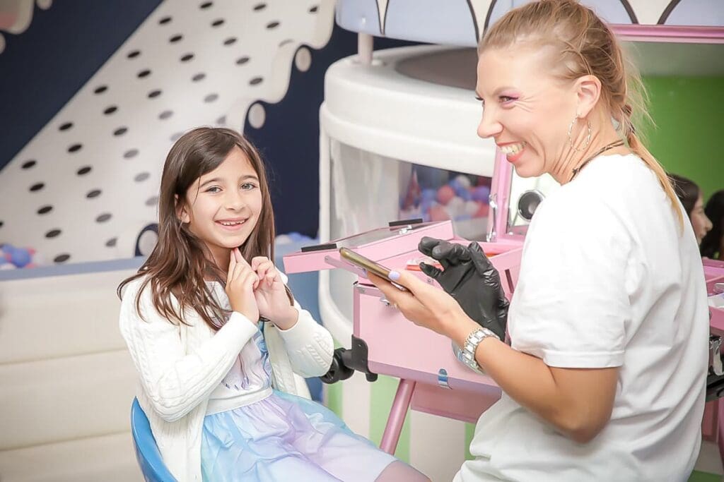 A young girl sits smiling in a chair while a woman wearing black gloves and holding a phone smiles beside her—a joyful moment captured by a kids birthday party photographer in a colorful, playful setting.