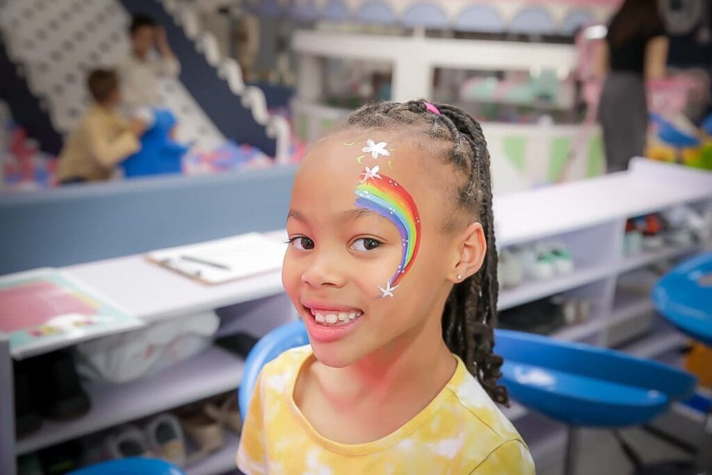 Smiling girl with braided hair and a colorful rainbow with stars painted on her face sits indoors, wearing a yellow tie-dye shirt. Captured by a skilled kids birthday party photographer, the background shows blurred shelves, blue chairs, and people.