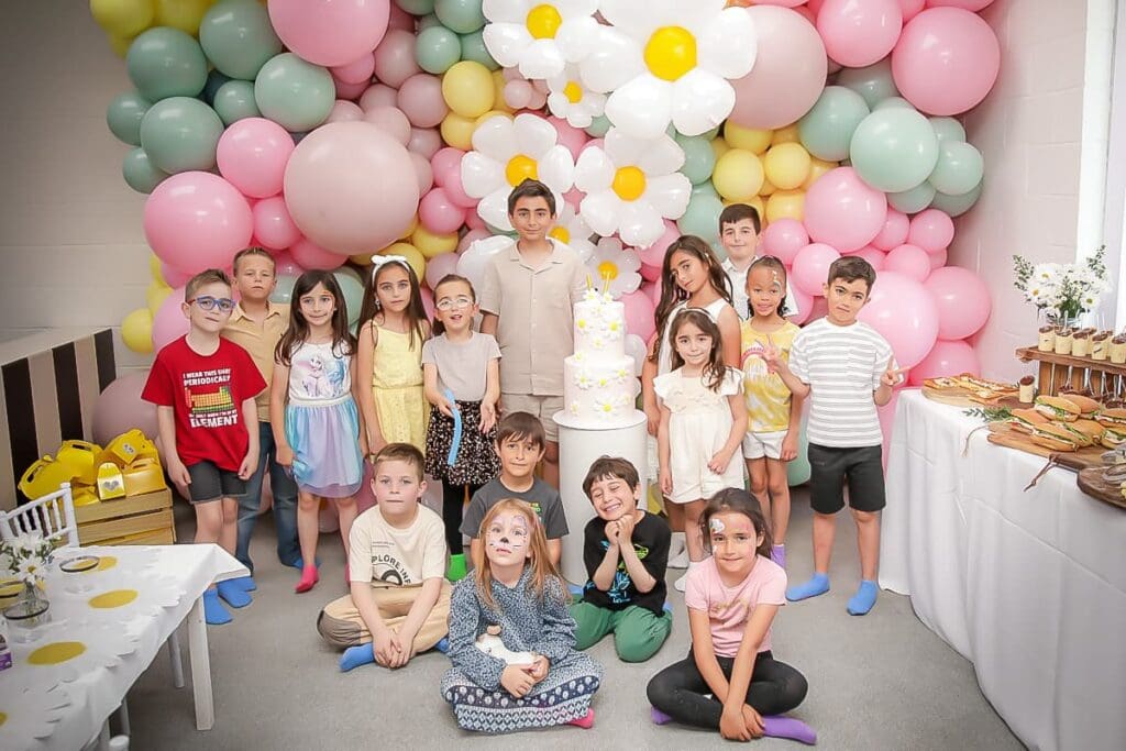 A group of children poses together at a birthday party with pastel balloons and a daisy cake. Some have face paint, all look cheerful, and festive decorations fill the scene—perfect for any kids birthday party photographer to capture.