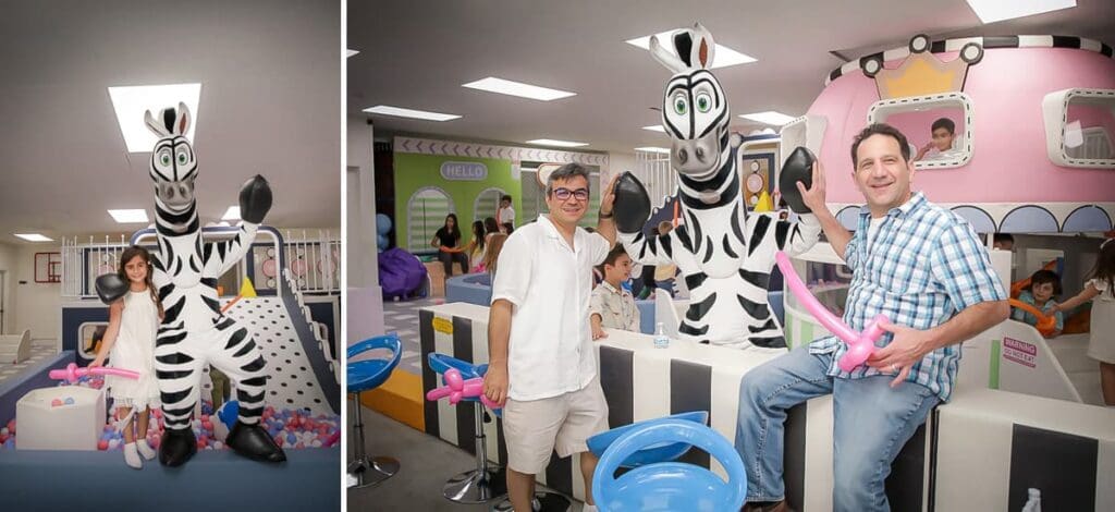 Two men, a woman, and children pose with a person in a zebra costume at an indoor play area. The men hold pink balloon animals as colorful play structures fill the background—perfect moments for a kids birthday party photographer to capture.