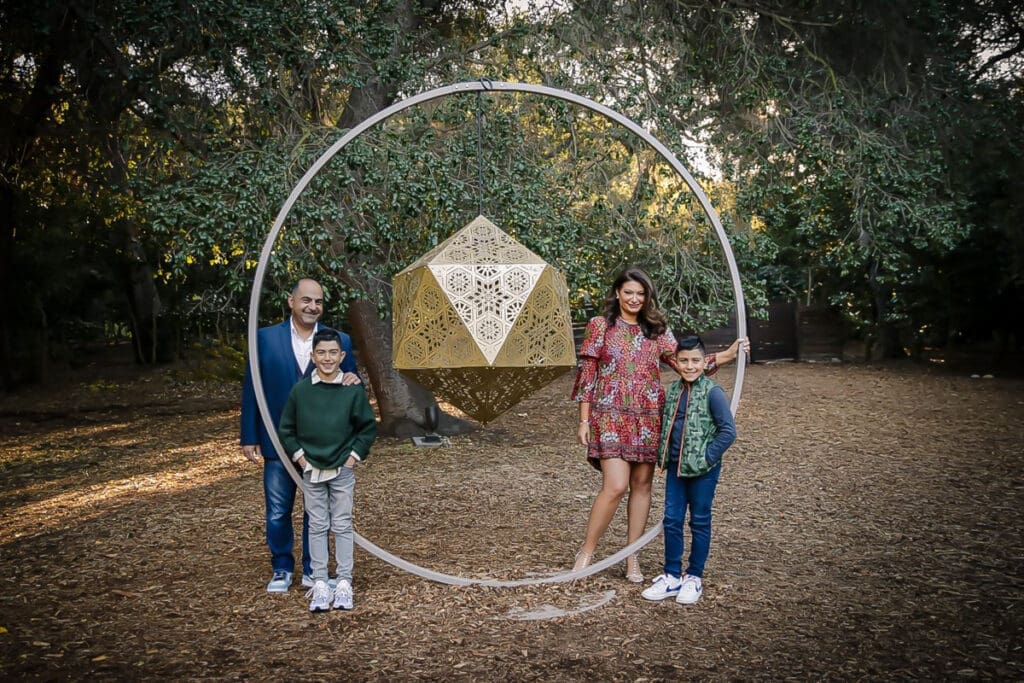 A family of four poses outdoors by a large geometric sculpture, trees and leaves in the background. Captured by a holiday family photographer, the parents stand behind their two young sons, all smiling in casual, colorful clothing.