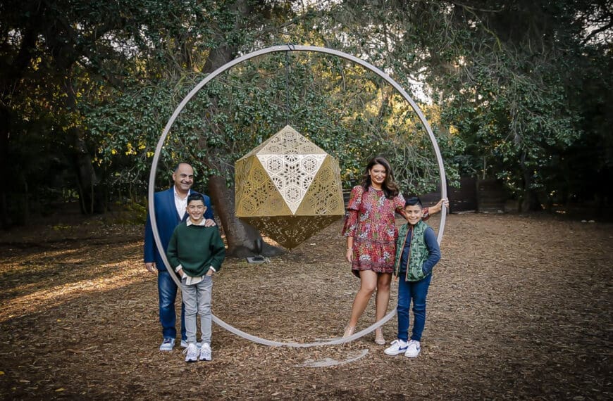 A family of four poses outdoors by a large geometric sculpture, trees and leaves in the background. Captured by a holiday family photographer, the parents stand behind their two young sons, all smiling in casual, colorful clothing.