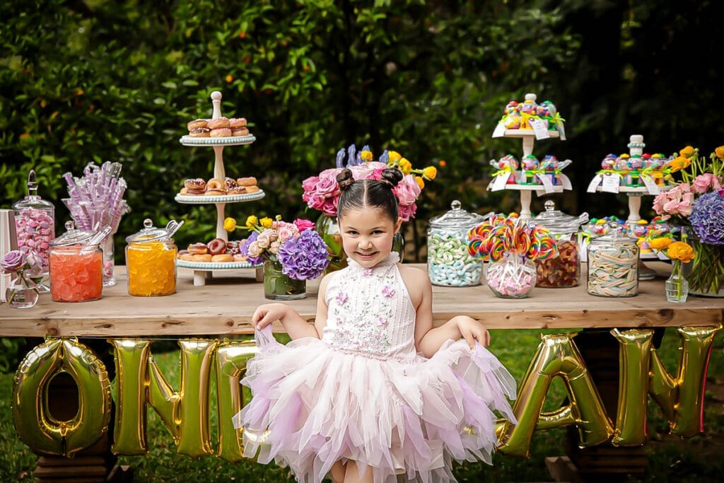 Smiling girl in a pink tutu by a colorful birthday candy and dessert table outdoors.