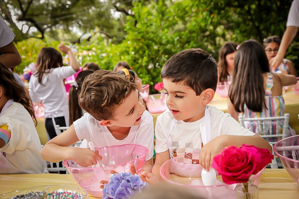 Two kids chat while mixing treats at an outdoor birthday party table with pink bowls and roses.