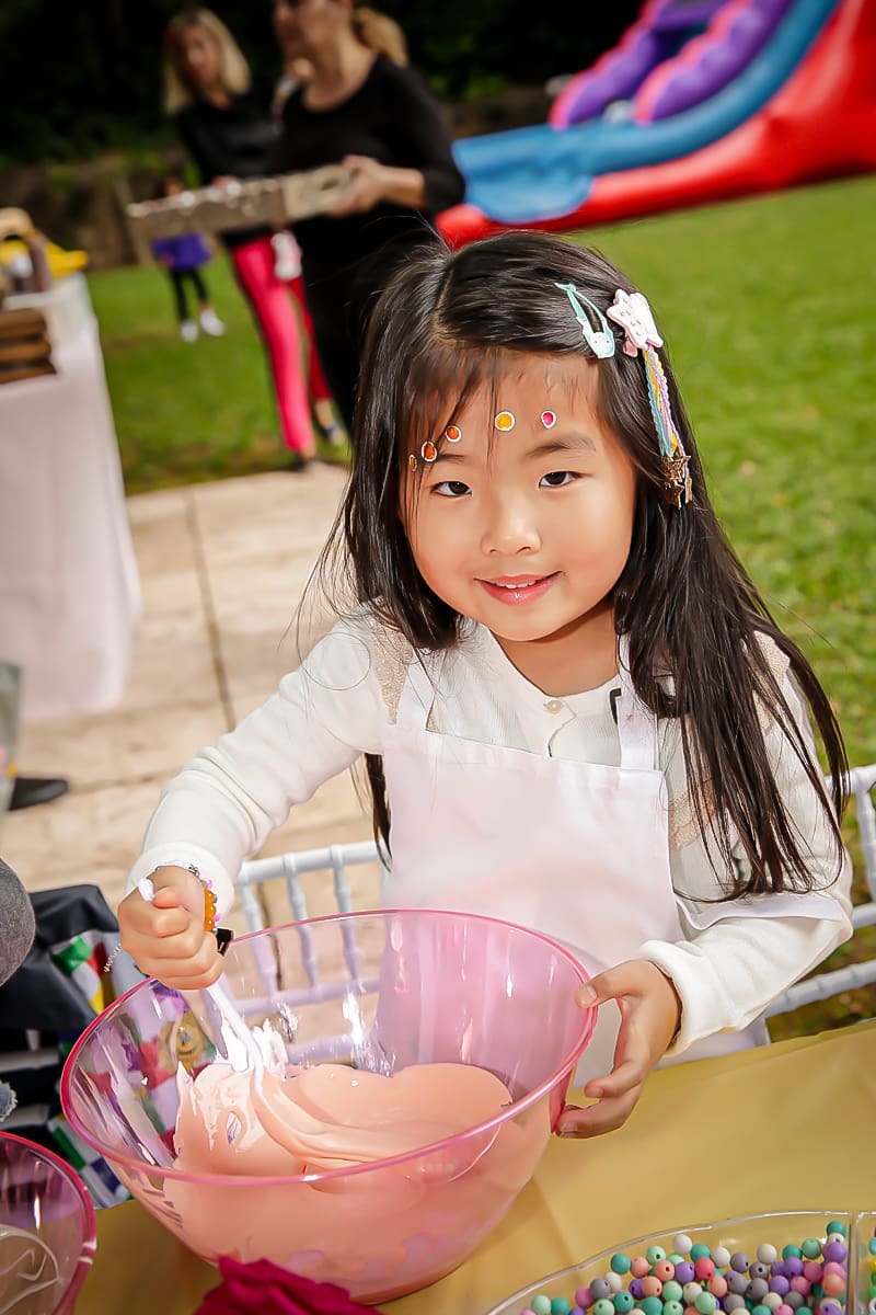 Smiling kid with stickers stirs pink slime at a backyard party.