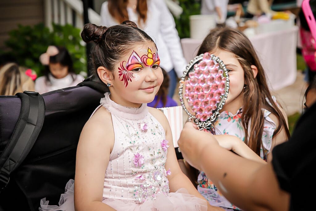 Smiling child with face paint admires a pink mirror at a birthday party.