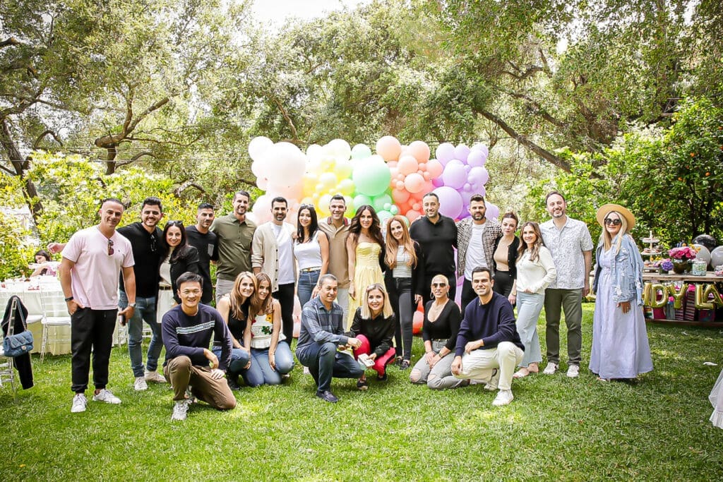 Group of adults posing at an outdoor birthday party with rainbow balloons.