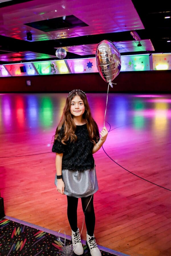Glendale 10th Birthday Party Photographer-Moonlight Rollerway A young girl with long brown hair, wearing a black top, silver skirt, black tights, and white boots, holds a silver balloon inside a colorful roller skating rink with wooden floors and bright lights.