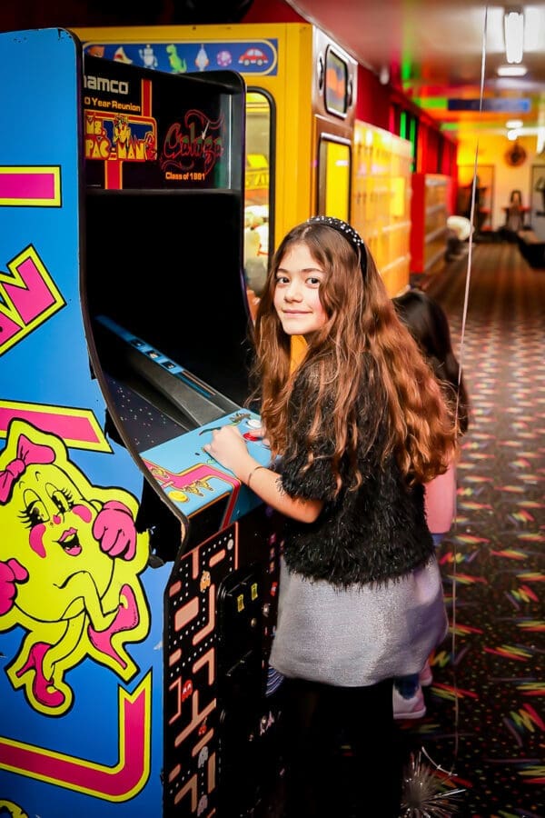 A young girl with long brown hair stands in front of a retro Ms. Pac-Man arcade machine in a brightly lit arcade, smiling at the camera.