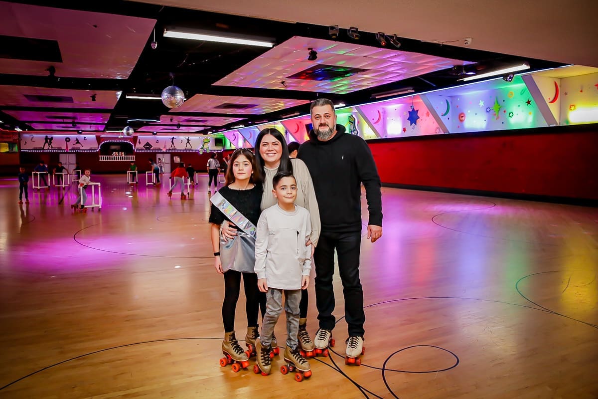 A family of four poses together on a brightly lit roller skating rink, all wearing roller skates. Colorful lights and decorations illuminate the background, creating a festive atmosphere.
