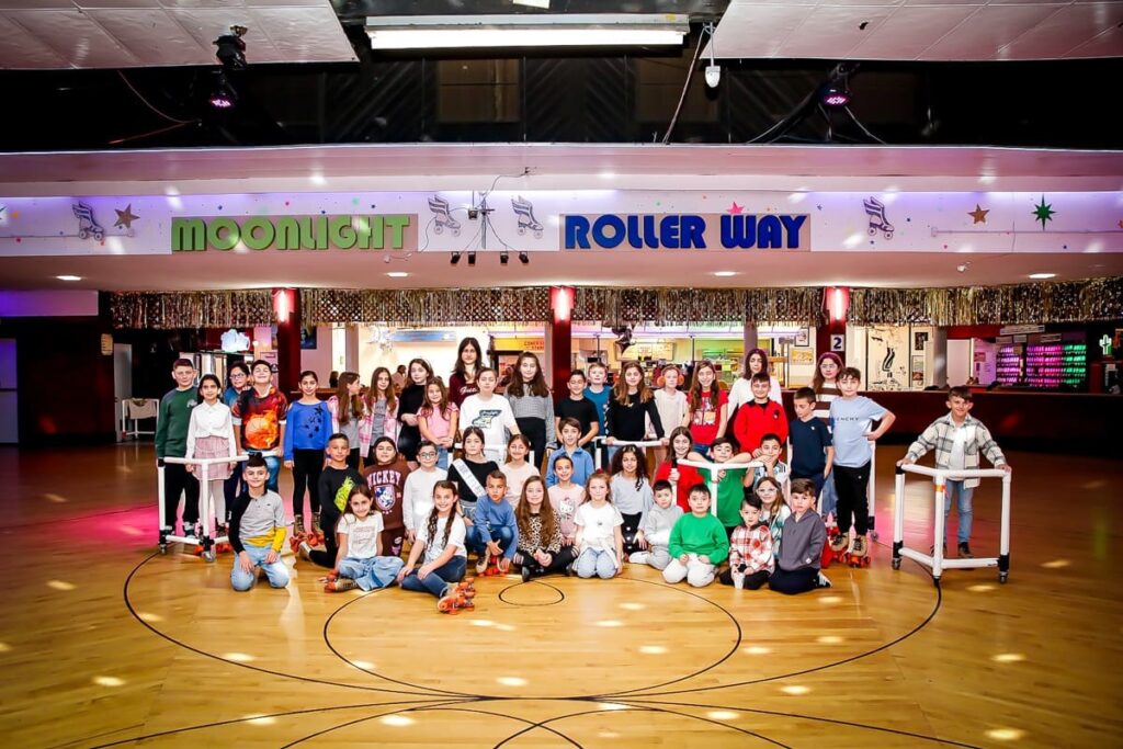 A large group of children and a few adults pose for a photo at an indoor roller skating rink called Moonlight Roller Way. Some are standing, while others sit or kneel on the wooden rink floor.