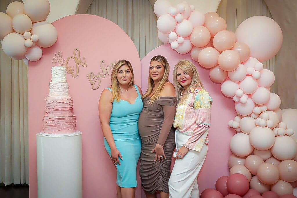 Three women pose in front of pink balloon decorations and a pink tiered cake. One woman is visibly pregnant, and all are dressed in stylish attire, smiling for the photo at what appears to be a baby shower.
