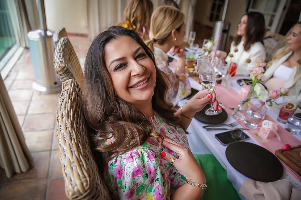 A woman in a floral dress smiles at the camera while holding a glass at a table set for a gathering with other women, decorated with pink flowers and tableware.