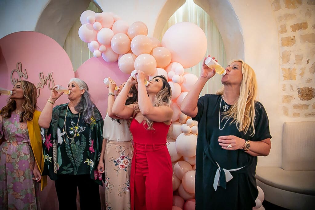 Five women stand in a row, tilting their heads back and drinking from small bottles, in front of a festive backdrop with pink and white balloons. They appear to be enjoying a celebration or party.