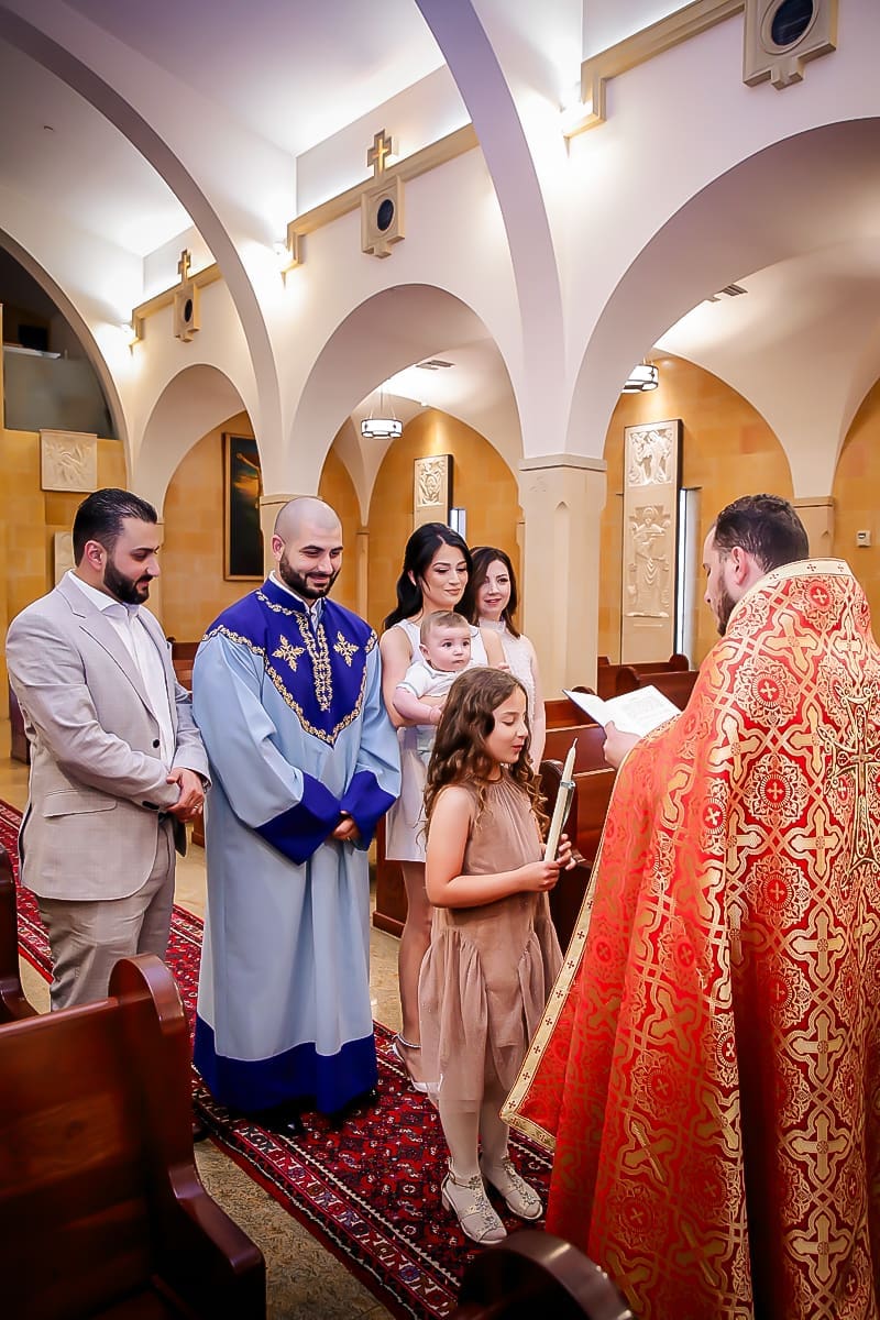 Family baptism ceremony; child holds a candle as priest reads.