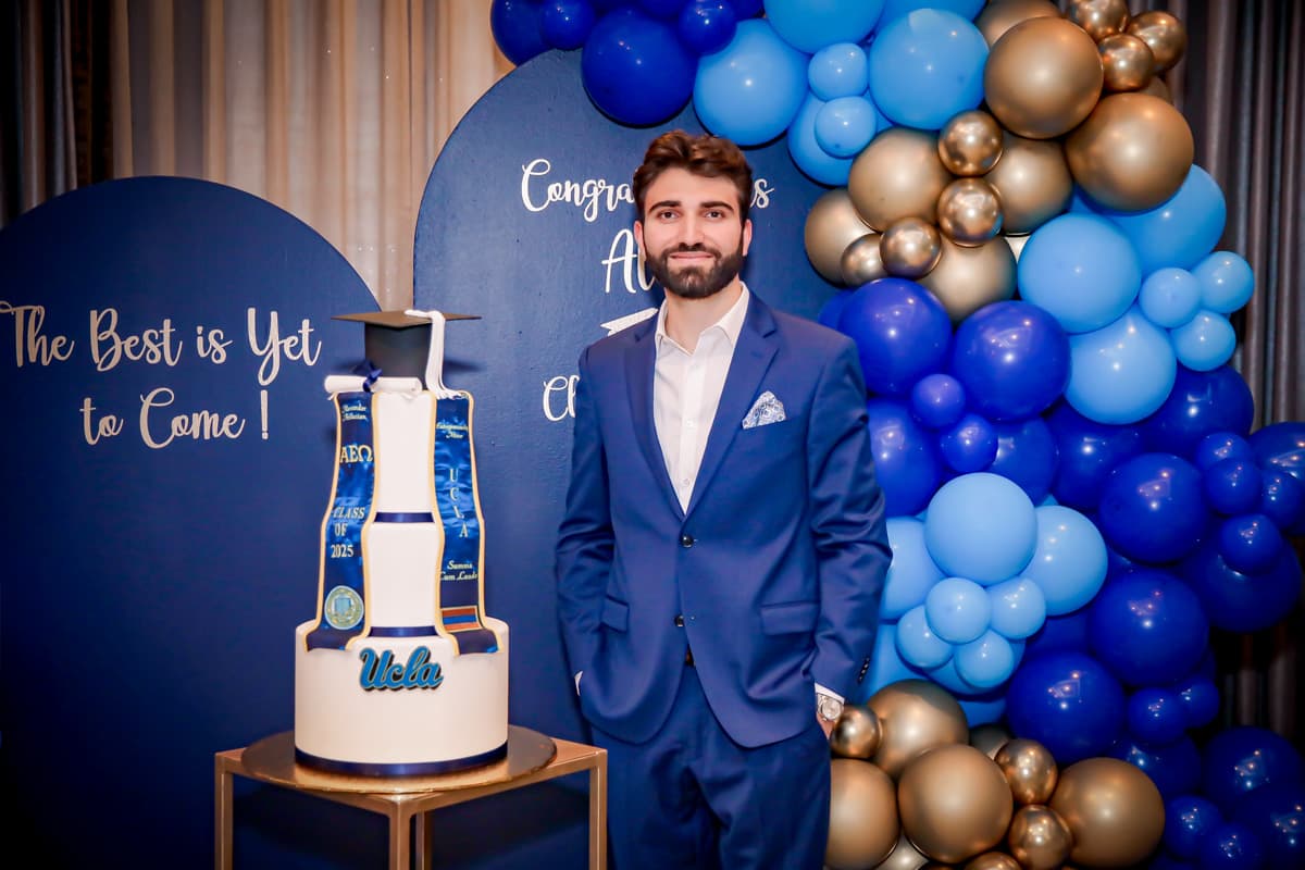 Young man dressed in formal attire in front of blue and gold decorations and balloons with a UCLA decorated graduation ckae. He he wearing a blue suit, smiling. Photography by Los Angeles Graduation Party Photographer, Jennifer Koochof